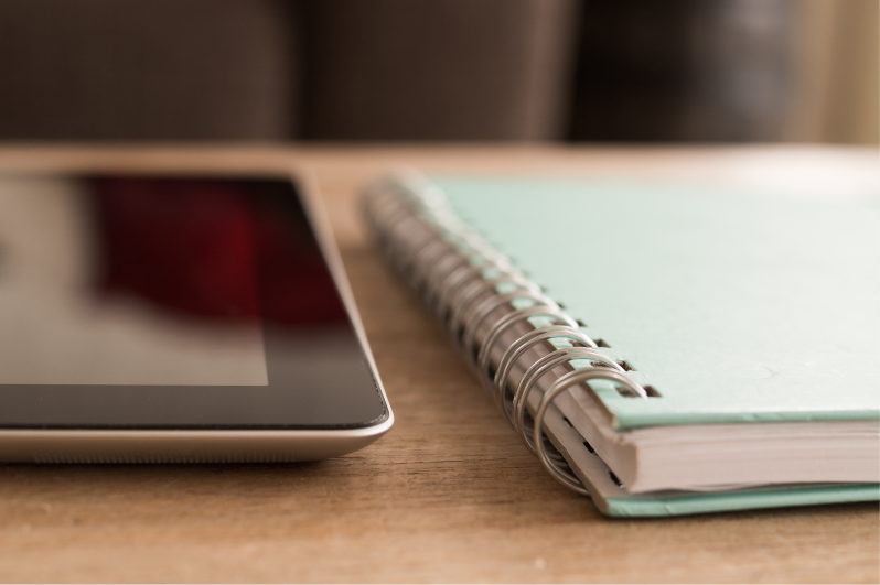 An electronic tablet and a spiral-bound notebook side by side on a table.